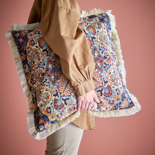 Person holding a decorative pillow with intricate patterns and fringes against a pink background