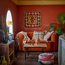 Load image into Gallery viewer, Living room with orange sofa, patterned cushions, and a colorful tapestry on a red wall.
