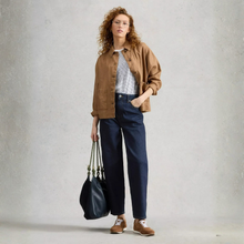 Load image into Gallery viewer, A woman stands against a textured light gray wall wearing a tan overshirt, white knit top, dark navy wide-leg trousers, and brown loafers, while holding a large black shoulder bag.
