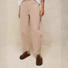Load image into Gallery viewer, A person wearing loose, straight-leg blush pink pants with decorative stitching down the sides, paired with a white shirt and dark brown suede mule loafers, standing against a neutral textured beige background.
