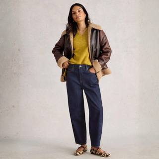 Woman wearing a brown leather jacket with fur trim, yellow sweater, and blue jeans against a plain background