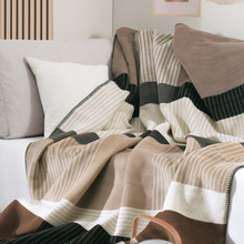 Load image into Gallery viewer, A cozy striped throw blanket in beige, cream, brown, and black tones draped over a light gray sofa with matching pillows in a softly lit living room.
