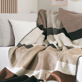 A cozy striped throw blanket in beige, cream, brown, and black tones draped over a light gray sofa with matching pillows in a softly lit living room.