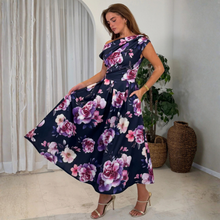 Load image into Gallery viewer, A woman models a navy blue off-the-shoulder maxi dress with a large pink and purple floral print. She is standing in a bright room with white curtains, a potted plant, and woven baskets.
