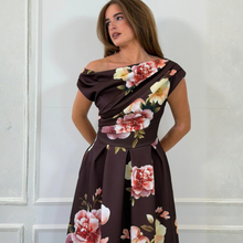 Load image into Gallery viewer, An up close look at the woman modeling a dark brown off-the-shoulder maxi dress with a large pink and white floral print, standing against a white paneled wall.
