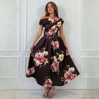A woman modeling a dark brown off-the-shoulder maxi dress with a large pink and white floral print, standing against a white paneled wall.