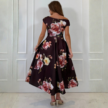 Load image into Gallery viewer, A woman wearing a dark brown off-the-shoulder midi dress with a large pink and white floral print, shown from the back against a white paneled wall.
