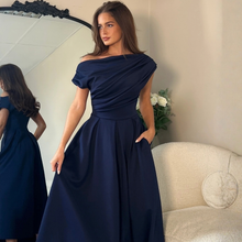 Load image into Gallery viewer, A woman models a navy blue off-the-shoulder maxi dress with a pleated bodice and skirt pockets, standing in a room with a cream-colored armchair and a mirror showing her back.

