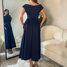 Load image into Gallery viewer, A woman wearing a navy blue off-the-shoulder maxi dress, shown from the back in a bright room with a cream-colored sofa and a mirror reflecting her profile.

