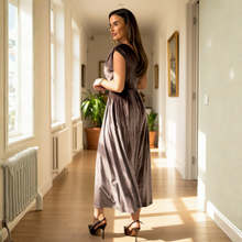 Load image into Gallery viewer, A woman in a sunlit hallway wearing a taupe-brown one-shoulder satin maxi dress with elegant draping, paired with black pointed-toe heels.

