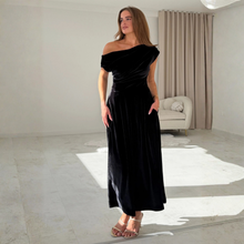 Load image into Gallery viewer, A woman modeling a black off-the-shoulder velvet maxi dress in a bright, modern room with beige walls, curtains, and a light-colored armchair.
