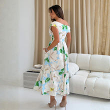 Load image into Gallery viewer, Woman in a floral dress standing in a living room with a white sofa and beige curtains.
