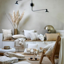 Load image into Gallery viewer, A styled living room corner with neutral-toned cushions, textured throws, a wooden chair, pampas grass in a vase, and a brass table lamp against a warm beige wall.
