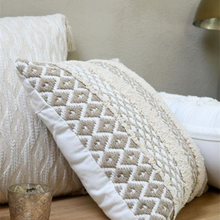 Load image into Gallery viewer, A decorative throw pillow with a cream and beige geometric diamond pattern and white piping, styled on a bed next to a textured cream pillow and a small glass candle holder.
