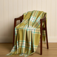 Load image into Gallery viewer, A woven plaid throw blanket in olive green, cream, and light blue is draped over a dark wooden chair against a cream paneled wall and light wood floor.
