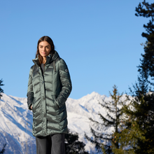 Load image into Gallery viewer, Woman wearing a green puffer coat standing in front of snow-capped mountains and trees.
