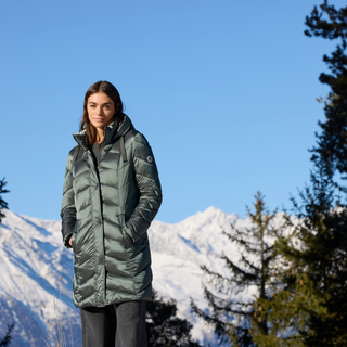Woman wearing a green puffer coat standing in front of snow-capped mountains and trees.