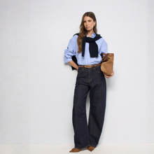 Load image into Gallery viewer, A model stands against a plain white studio background wearing a light blue button-up shirt, dark wide-leg jeans, a black knit sweater draped around her shoulders, brown suede boots, and holding a brown suede bag.
