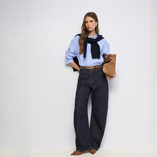 A model stands against a plain white studio background wearing a light blue button-up shirt, dark wide-leg jeans, a black knit sweater draped around her shoulders, brown suede boots, and holding a brown suede bag.