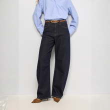 Load image into Gallery viewer, A person wearing dark wide-leg denim jeans with a brown belt, paired with a light blue button-up shirt and brown pointed flats, standing against a plain light gray wall.
