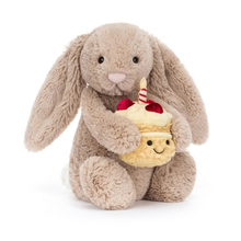 Load image into Gallery viewer, A plush toy bunny in warm taupe holding a small yellow cupcake character with a smiling face, red cherries, and a striped candle, photographed on a clean white background.

