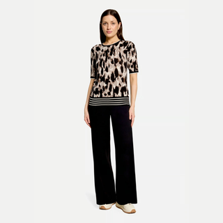 A woman modeling a short-sleeve knit top with a beige, brown, and black leopard print and horizontal black stripes at the hem, paired with black wide-leg trousers, against a plain white background.
