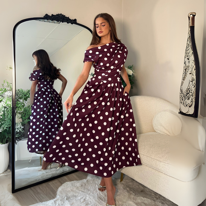 Woman in a purple polka dot dress standing in front of a mirror in a room with a white armchair and decorative items.
