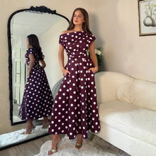 Load image into Gallery viewer, A woman stands in a living room wearing a burgundy off-the-shoulder maxi dress with white polka dots. She is posed in front of a large black-framed mirror that reflects her side profile, next to a cream-colored sofa.
