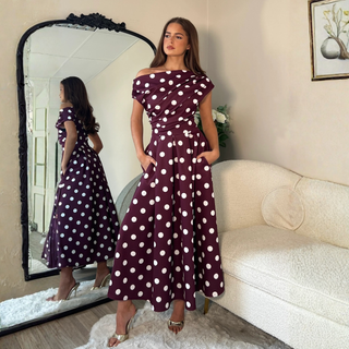 A woman stands in a living room wearing a burgundy off-the-shoulder maxi dress with white polka dots. She is posed in front of a large black-framed mirror that reflects her side profile, next to a cream-colored sofa.