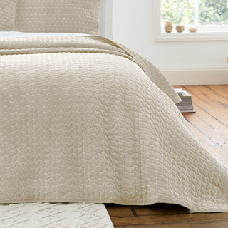 Beige quilted coverlet on a bed in a room with a window and wooden floor.
