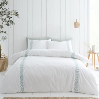 White bedding set with decorative trim on a bed in a bedroom with a wooden floor and plant.