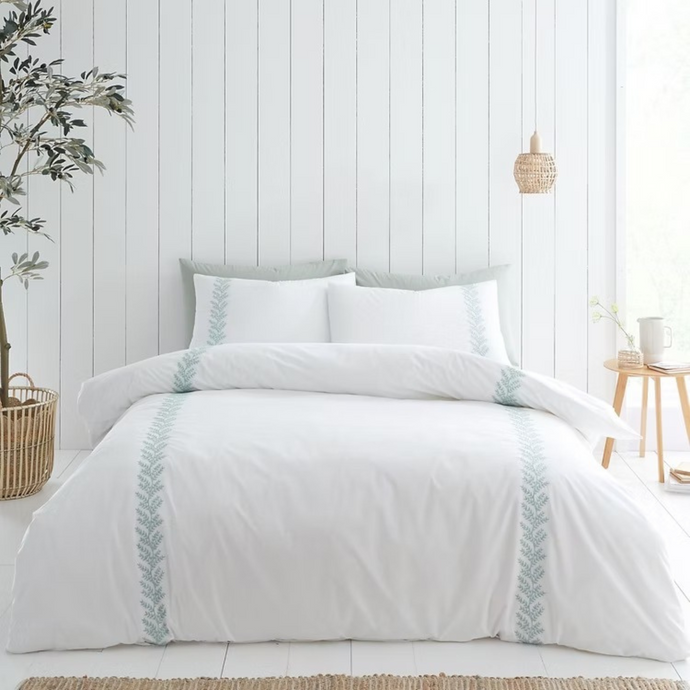 White bedding set with decorative trim on a bed in a bedroom with a wooden floor and plant.