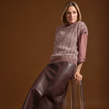 Load image into Gallery viewer, A woman models a knit sweater vest with a geometric pattern over a striped long-sleeve shirt, paired with a dark brown faux-leather skirt, posed against a solid brown studio background.
