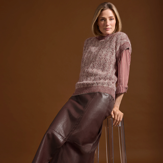A woman models a knit sweater vest with a geometric pattern over a striped long-sleeve shirt, paired with a dark brown faux-leather skirt, posed against a solid brown studio background.