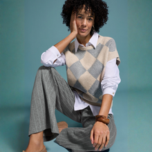 Load image into Gallery viewer, A woman poses in a studio setting wearing a beige and gray argyle knit sweater vest over a white collared shirt, paired with gray pinstriped trousers, against a solid teal background.
