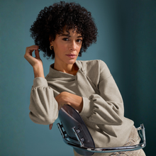 Load image into Gallery viewer, A woman with curly dark hair wearing a light beige long-sleeve knit top and patterned trousers, holding a dark blue clutch bag while seated on a chrome chair against a solid teal background.
