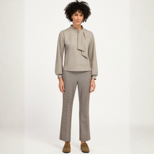 Load image into Gallery viewer, A model stands against a plain light background wearing a light taupe long-sleeve blouse with a bow detail at the collar and matching plaid trousers in beige, brown, and blue tones.
