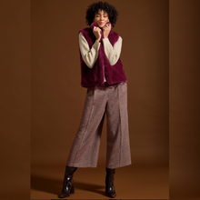 Load image into Gallery viewer, A model stands against a solid brown studio backdrop wearing a deep burgundy faux-fur vest over a cream ribbed sweater, paired with wide-leg taupe tweed trousers and black ankle boots.
