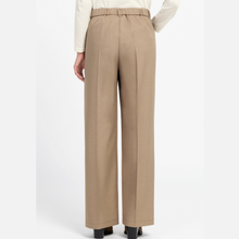 Load image into Gallery viewer, A person shown from the back wearing high-waisted, wide-leg trousers in a warm beige/tan color, paired with a cream ribbed long-sleeve top and black shoes, against a plain light gray studio background.
