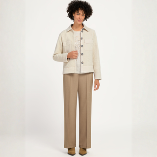A model stands against a plain white studio background wearing a textured cream jacket over a white knit top, paired with straight-leg beige trousers and brown shoes.