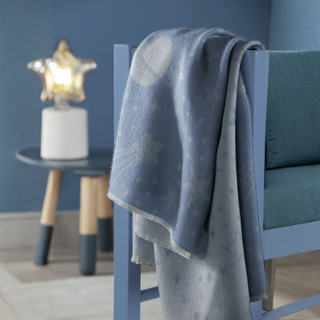 A light blue knit blanket with a white moon and star pattern draped over a blue wooden chair in a styled room setting.