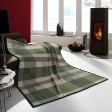 Load image into Gallery viewer, A plaid throw blanket in shades of green, beige, and gray is draped over a dark sofa in a modern living room setting with a fireplace and large windows.
