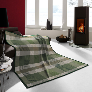 A plaid throw blanket in shades of green, beige, and gray is draped over a dark sofa in a modern living room setting with a fireplace and large windows.