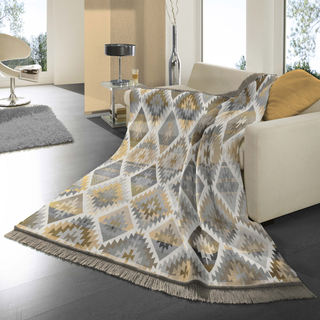 A patterned throw blanket draped over a cream sofa in a modern living room. The blanket features a geometric diamond design in beige, gray, and white with fringed edges.