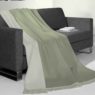 A sage green and off-white textured throw blanket with fringe is draped over a dark gray fabric sofa in a modern room setting.