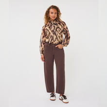 Load image into Gallery viewer, A model stands against a light gray studio background wearing a long-sleeve leopard-print blouse with a high neck and wide-leg brown trousers, paired with black and white sneakers.
