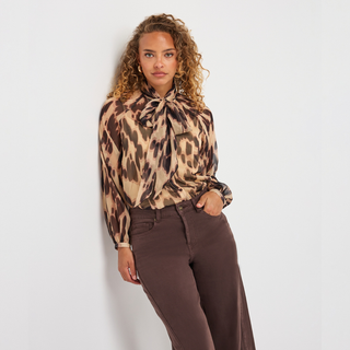 A woman with curly hair models a long-sleeve leopard print blouse with a tie-neck detail, paired with dark brown wide-leg pants, against a plain white studio background.