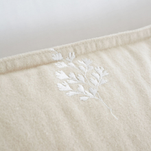 Load image into Gallery viewer, Close-up of a cream-colored textured fabric item, likely a towel or blanket, featuring a white embroidered leafy branch motif and a stitched border.
