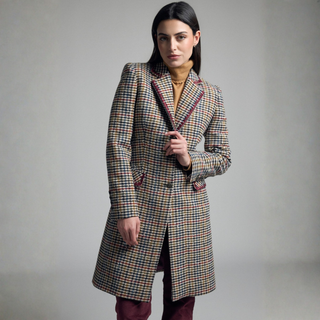 A woman modeling a mid-length plaid coat with a beige base and multicolor check pattern, paired with dark trousers and a mustard top, against a neutral gray studio background.