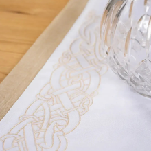 Load image into Gallery viewer, Close-up of a white tablecloth with gold embroidery and a clear glass on a wooden surface.
